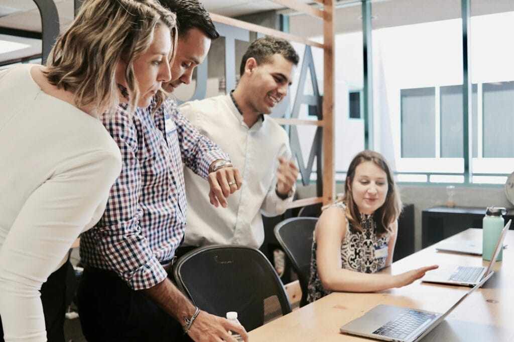 Four people from the web development team gather around a table, engaged in a discussion while looking at a laptop screen in an office setting. on possibleweb.com.